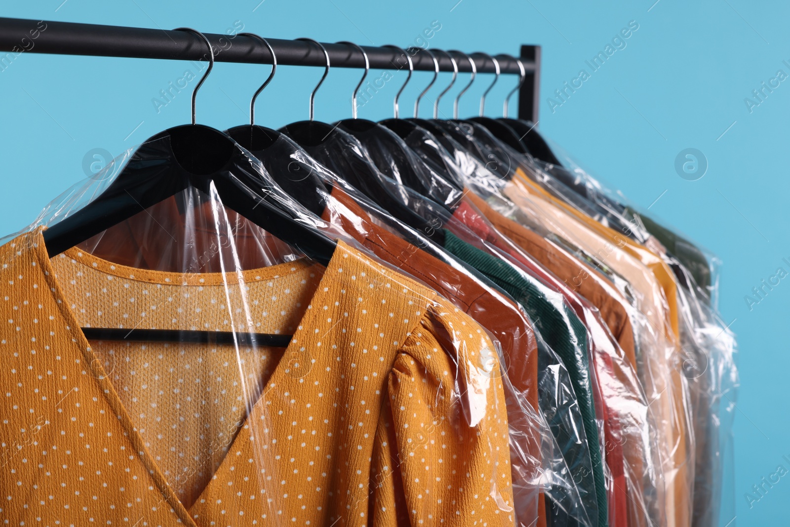 Photo of Dry-cleaning service. Many different clothes in plastic bags hanging on rack against light blue background