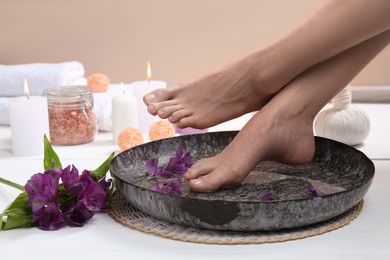 Photo of Woman holding her feet over bowl with water and flowers on white wooden floor, closeup. Spa treatment