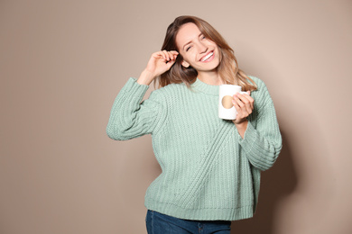 Happy young woman in warm sweater with cup of hot drink on beige background