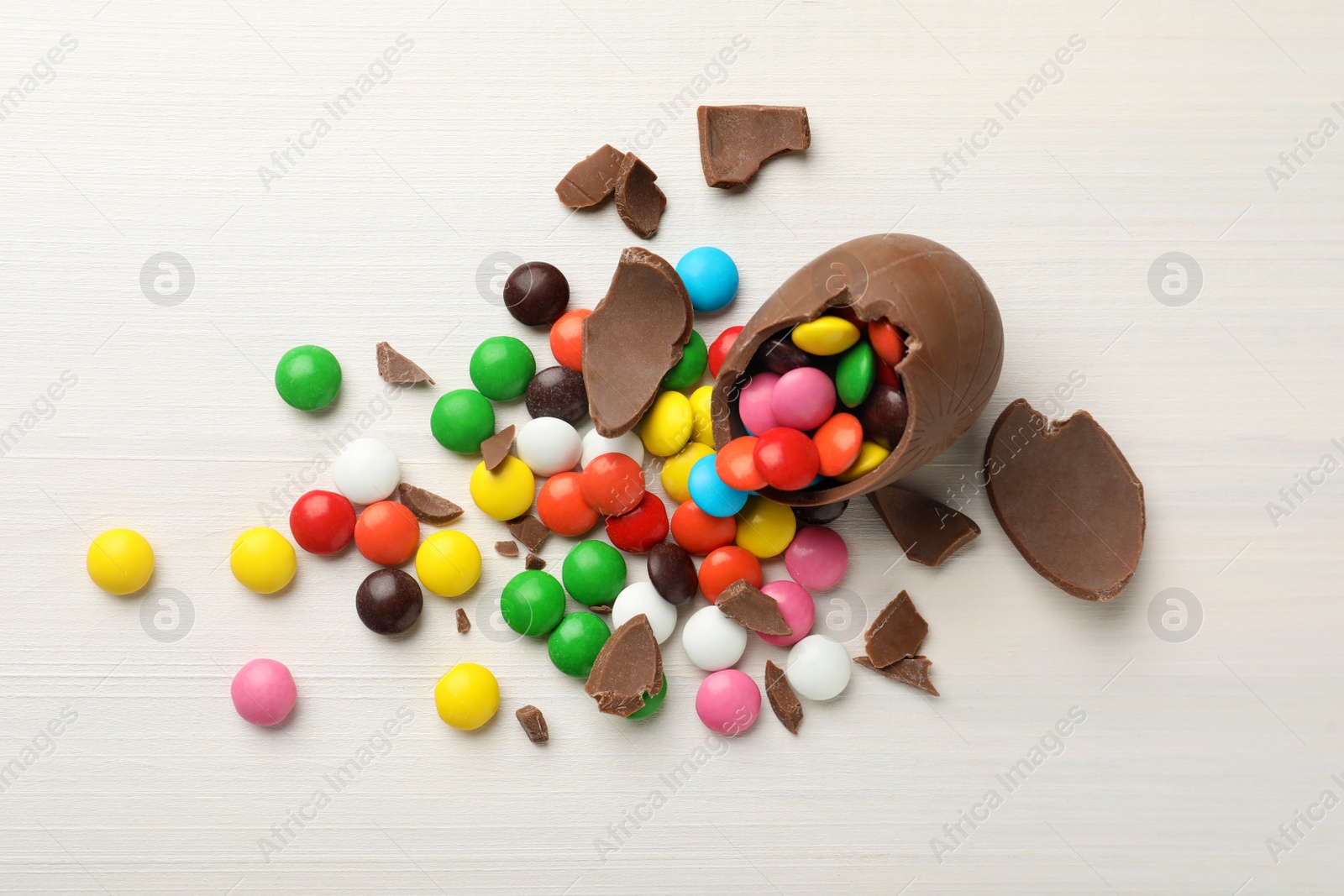 Photo of Broken chocolate egg and colorful candies on white wooden table, flat lay