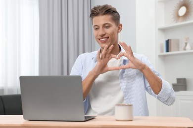 Photo of Happy young man having video chat via laptop and making heart at wooden table indoors. Long-distance relationship