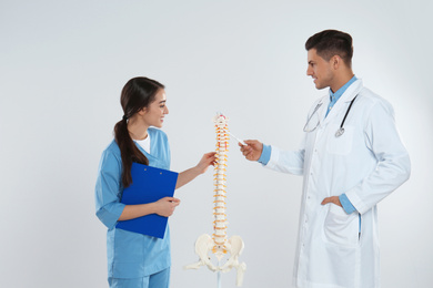 Professional orthopedist with human spine model teaching medical student against light background