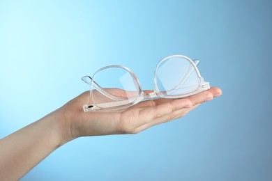 Woman holding glasses with transparent frame on light blue background, closeup
