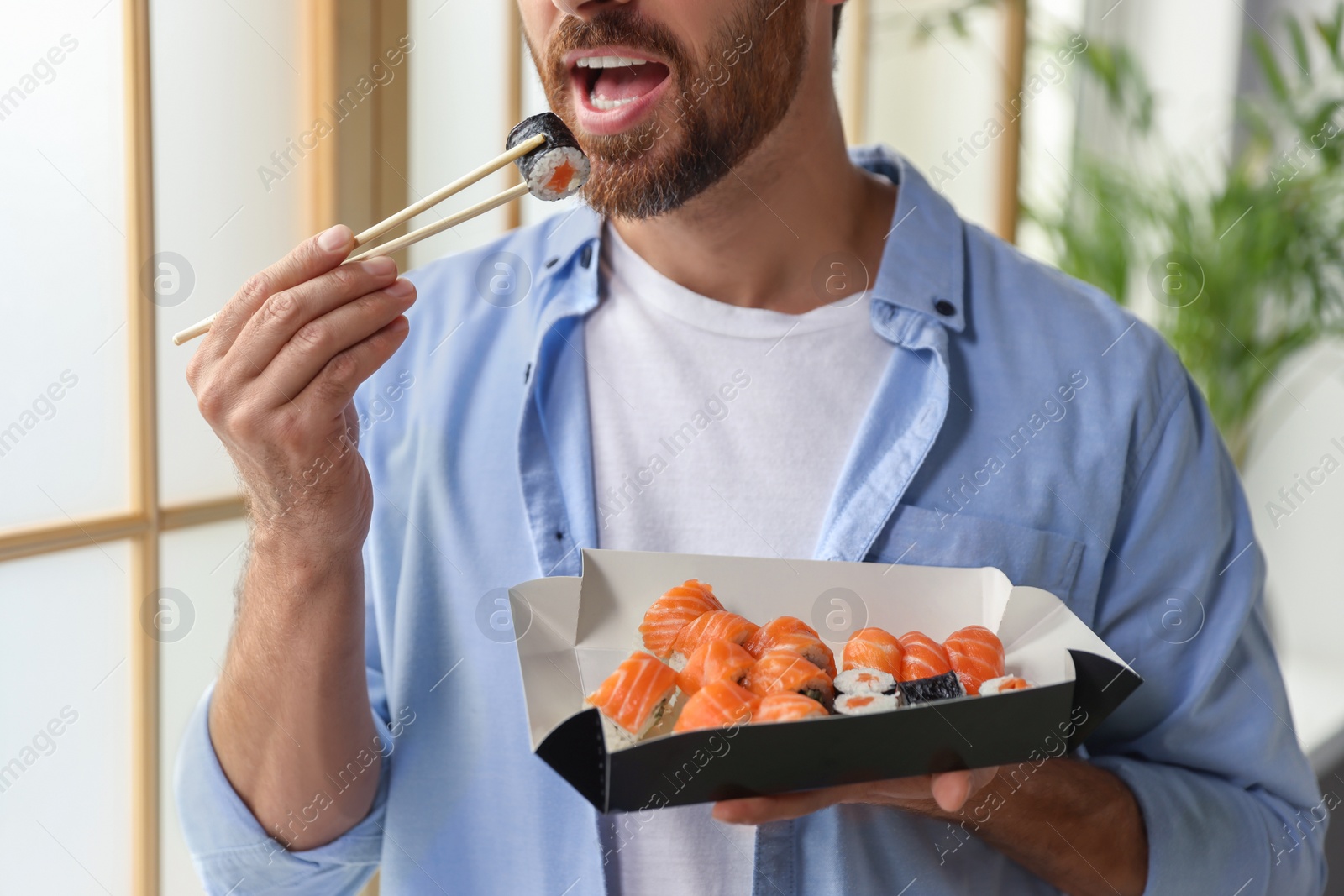 Photo of Handsome man eating sushi rolls with chopsticks indoors, closeup