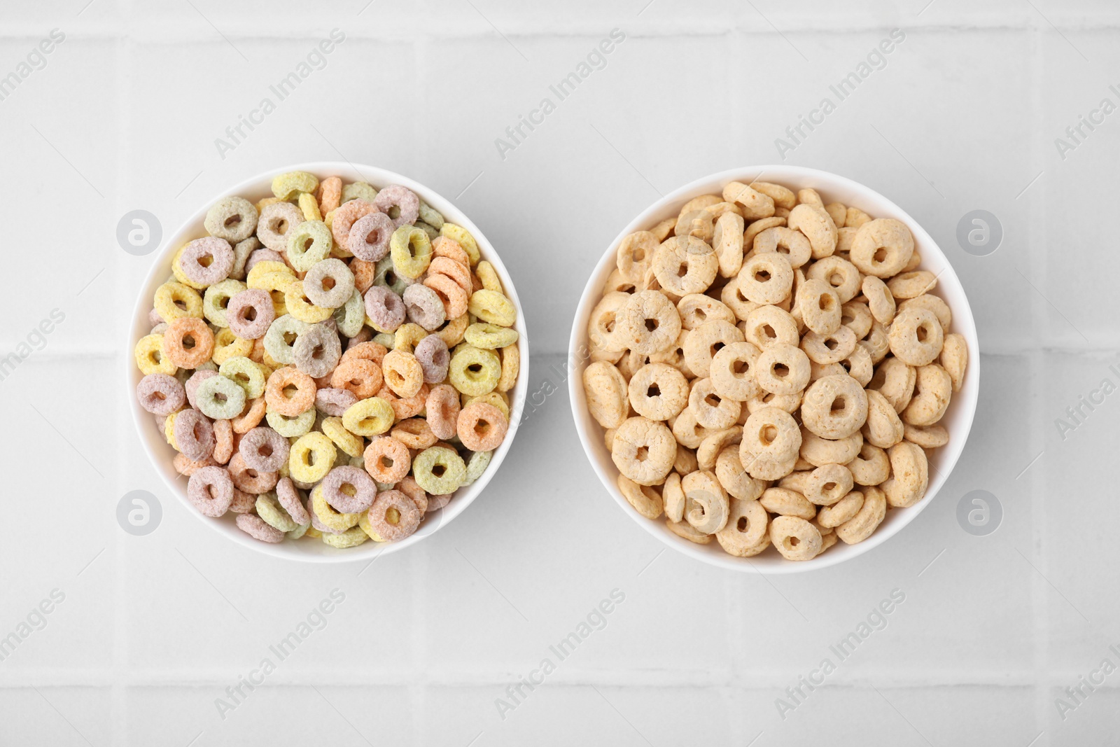 Photo of Tasty cereal rings in bowls on white tiled table, flat lay