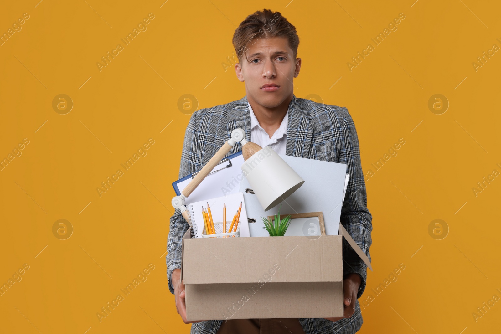 Photo of Unemployed young man with box of personal office belongings on orange background