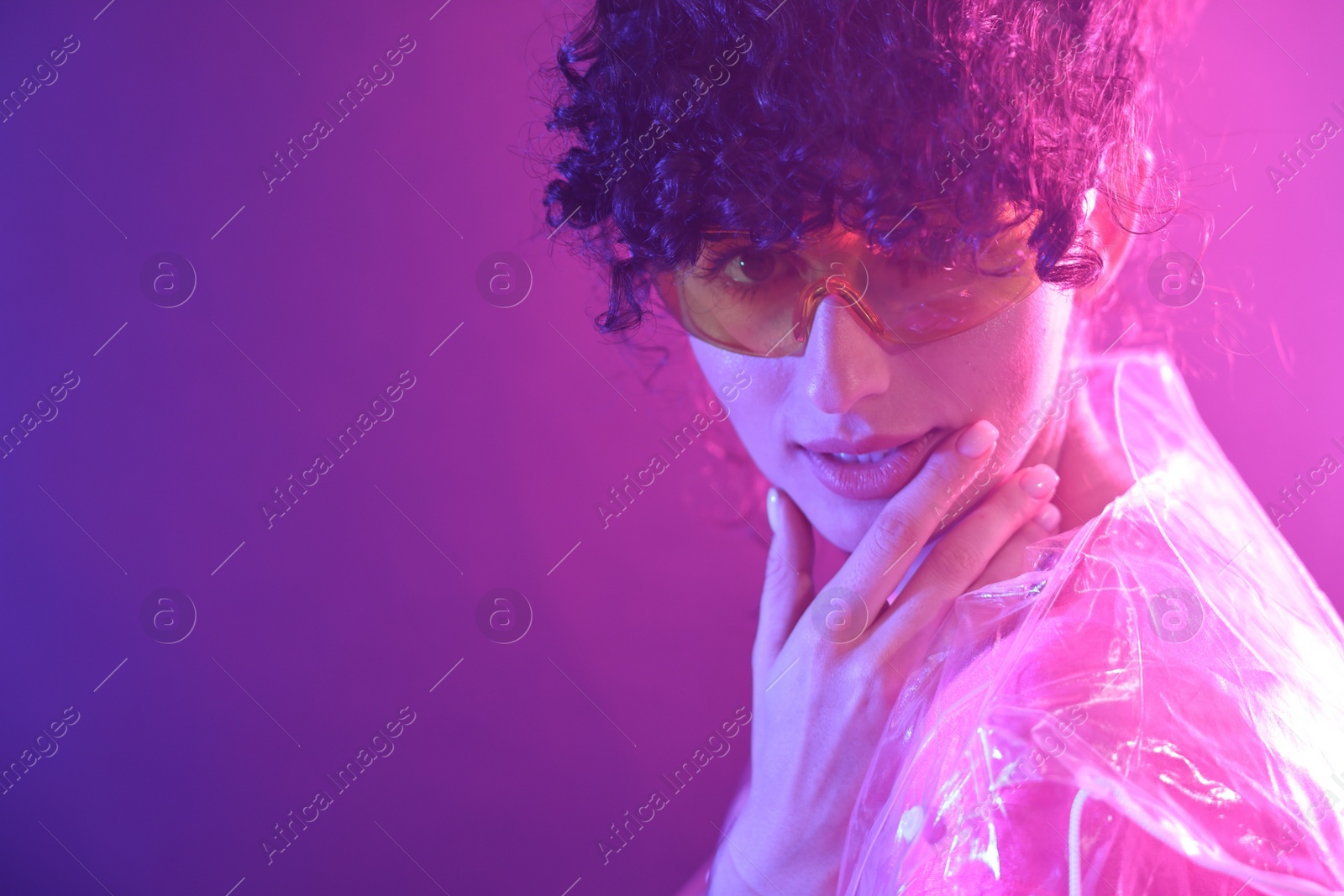 Photo of Beautiful young woman in transparent coat and sunglasses posing on color background in neon lights