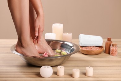 Photo of Woman soaking her feet in bowl with water and flowers on wooden surface, closeup. Pedicure procedure