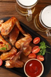 Mugs with beer, delicious baked chicken wings and sauce on wooden table, flat lay