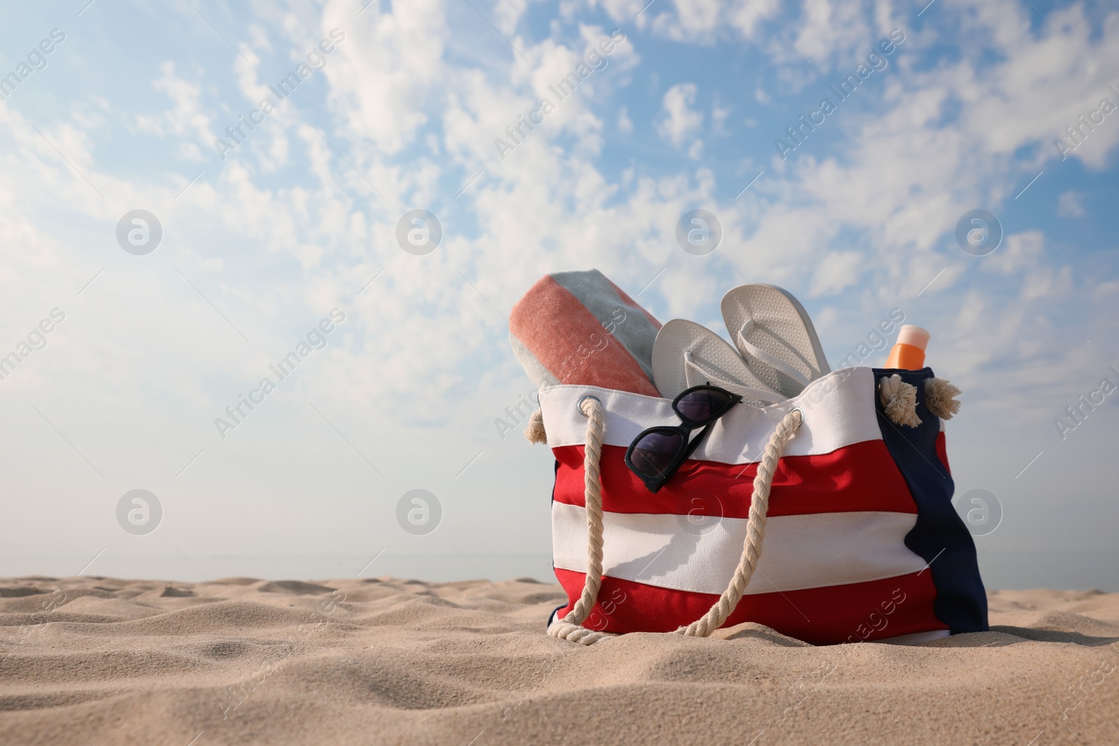 Photo of Beach bag with flip flops, towel, sunglasses and sunscreen on sand, space for text