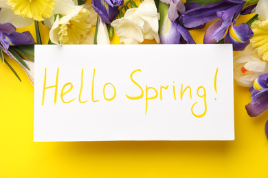 Photo of Card with words HELLO SPRING and fresh flowers on yellow background, flat lay