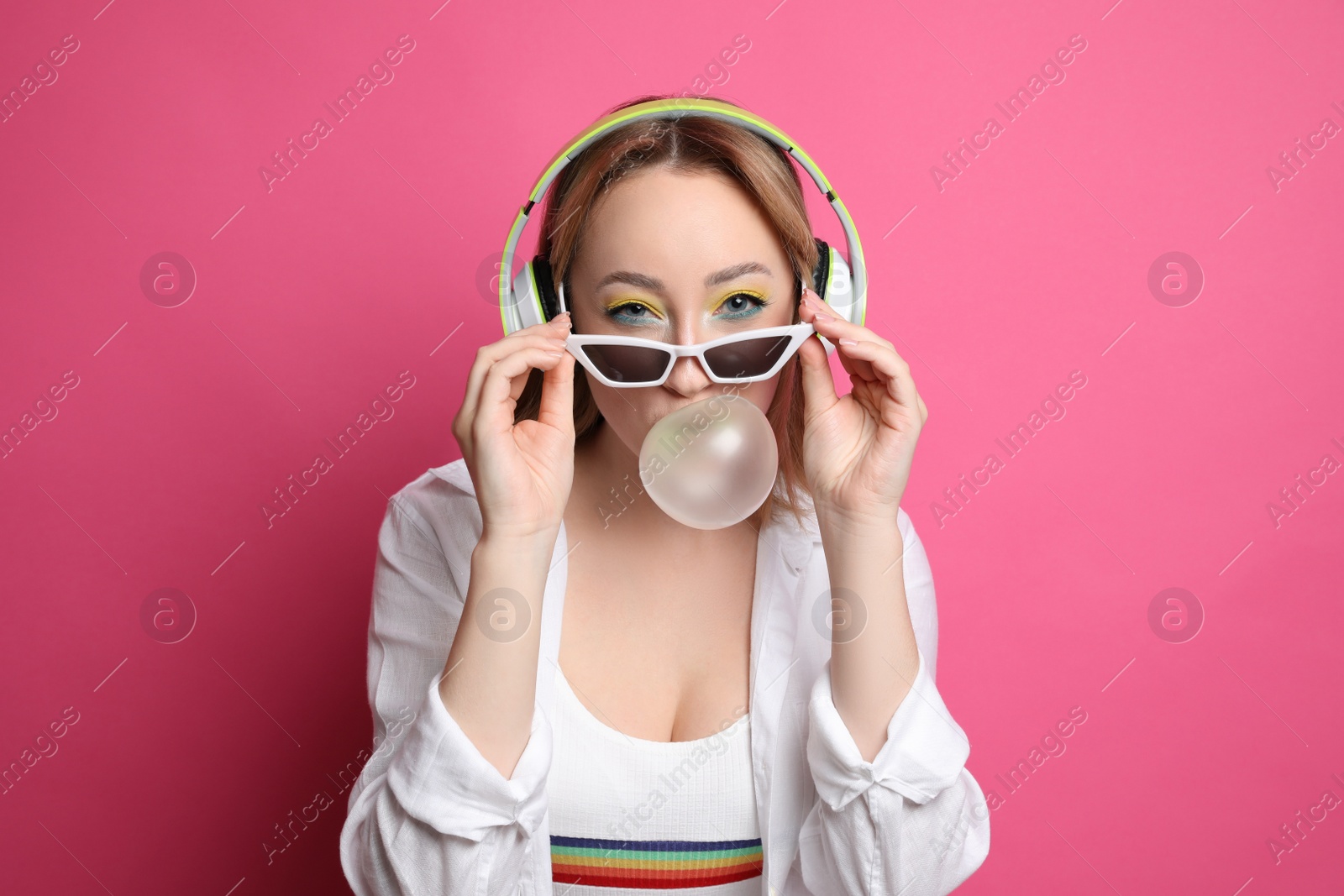 Photo of Fashionable young woman with bright makeup and headphones blowing bubblegum on pink background