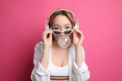 Fashionable young woman with bright makeup and headphones blowing bubblegum on pink background