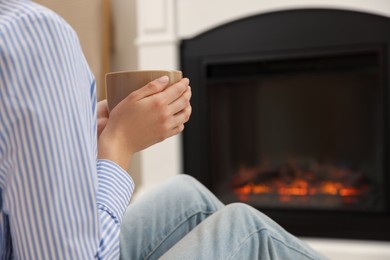 Photo of Young woman with cup of hot drink resting near fireplace at home, closeup