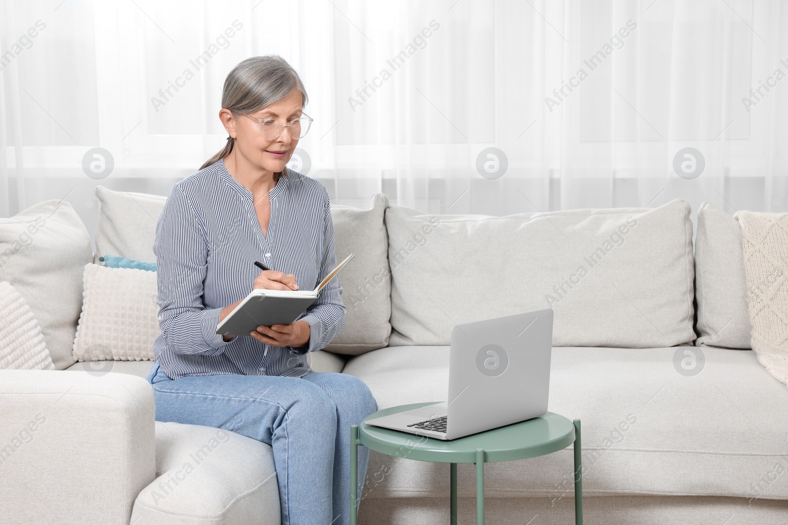 Photo of Beautiful senior woman writing something in notebook while working with laptop at home