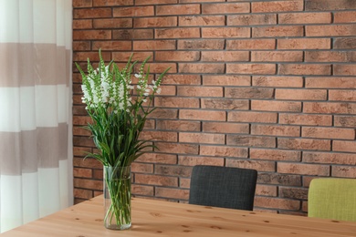 Vase with beautiful flowers on wooden table against brick wall. Stylish interior