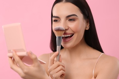 Beautiful woman with cosmetic pocket mirror applying makeup on pink background