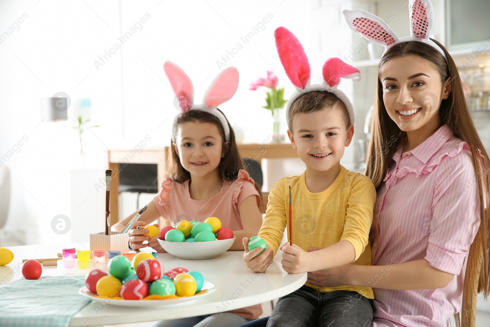 Photo of Mother and her children with bunny ears headbands painting Easter eggs in kitchen, space for text
