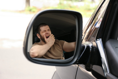 Photo of Tired man yawning in his auto, view through car side mirror