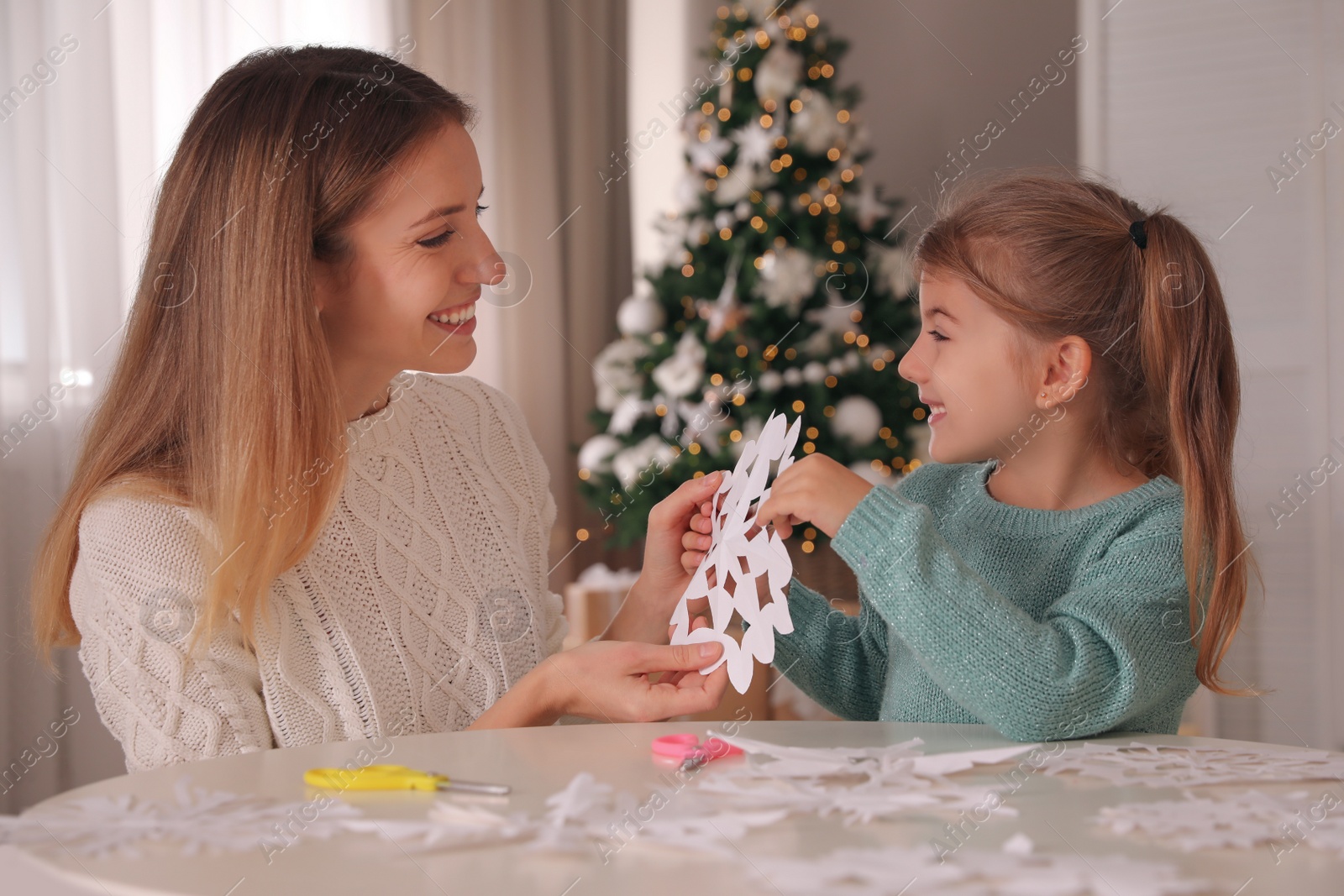 Photo of Happy mother and daughter making paper snowflake at table near Christmas tree indoors