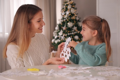 Happy mother and daughter making paper snowflake at table near Christmas tree indoors