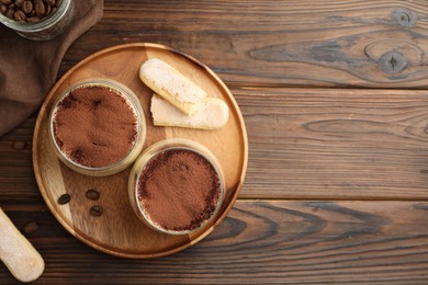 Photo of Delicious tiramisu in glasses, coffee beans and cookies on wooden table, flat lay. Space for text