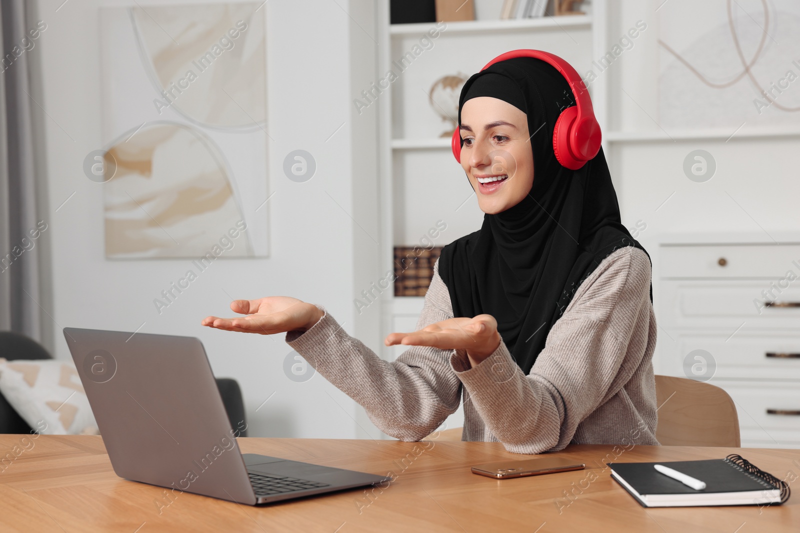 Photo of Muslim woman in hijab using video chat on laptop at wooden table in room