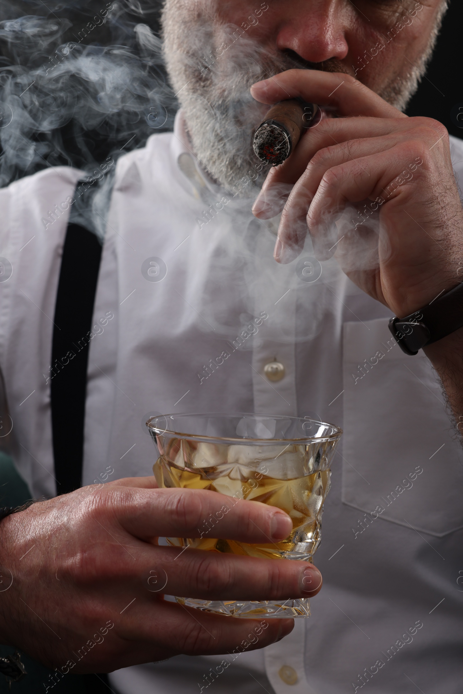 Photo of Bearded man with glass of whiskey smoking cigar against black background, closeup