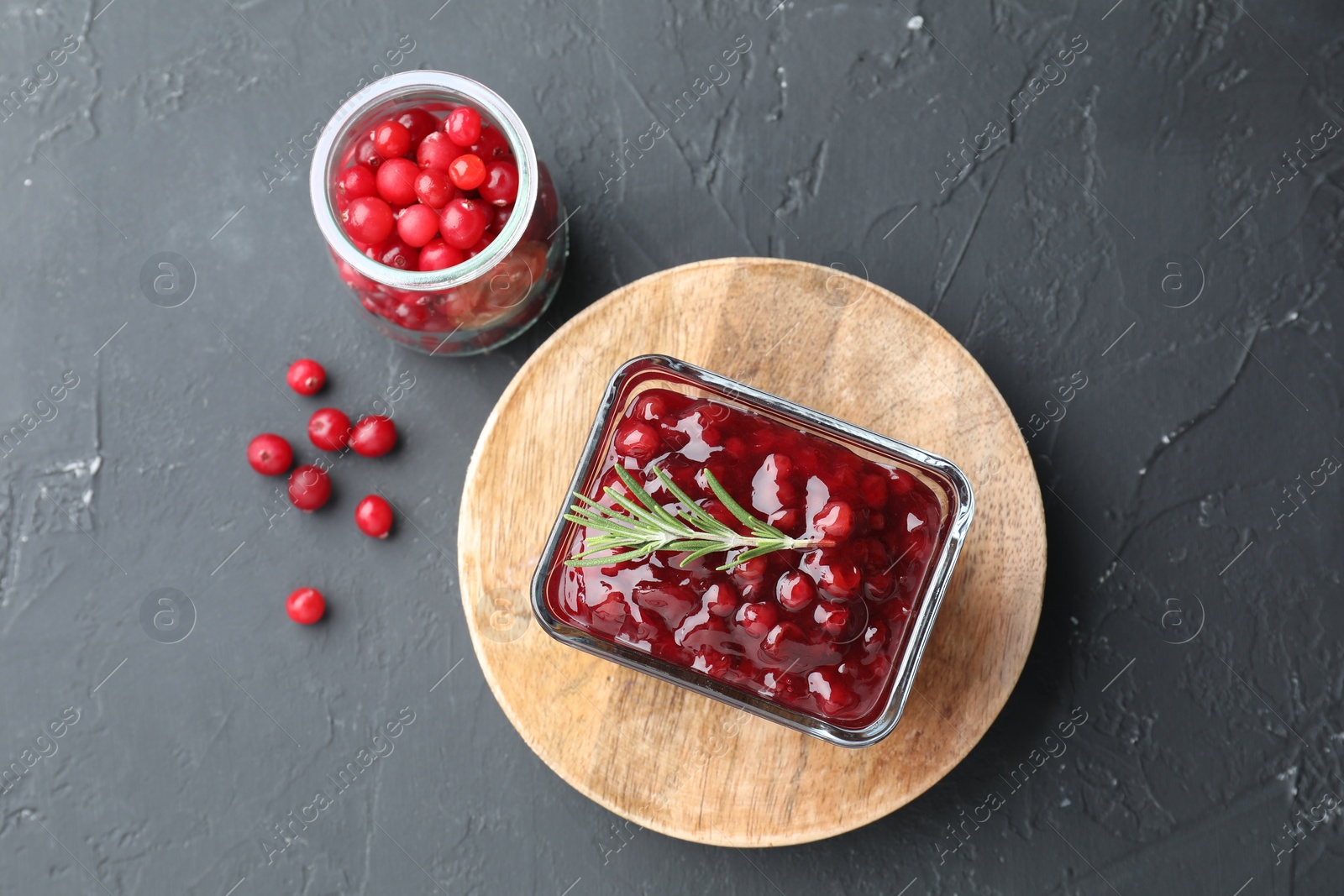 Photo of Tasty cranberry sauce in bowl and glass jar with fresh berries on gray textured table, flat lay