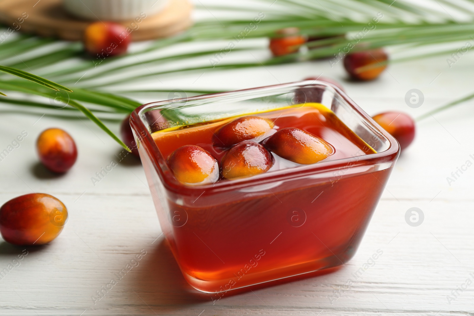 Image of Palm oil in glass bowl with fruits and tropical leaves on white wooden table, closeup