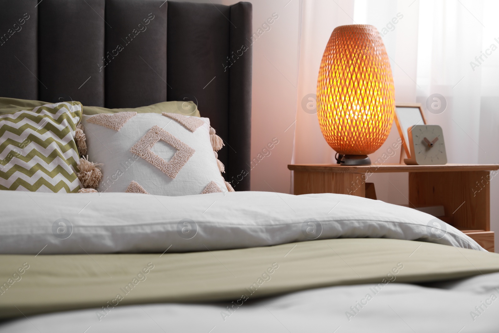 Photo of Comfortable bed with cushions, lamp and different decor on wooden bedside table in room. Stylish interior