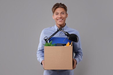 Happy unemployed young man with box of personal office belongings on grey background