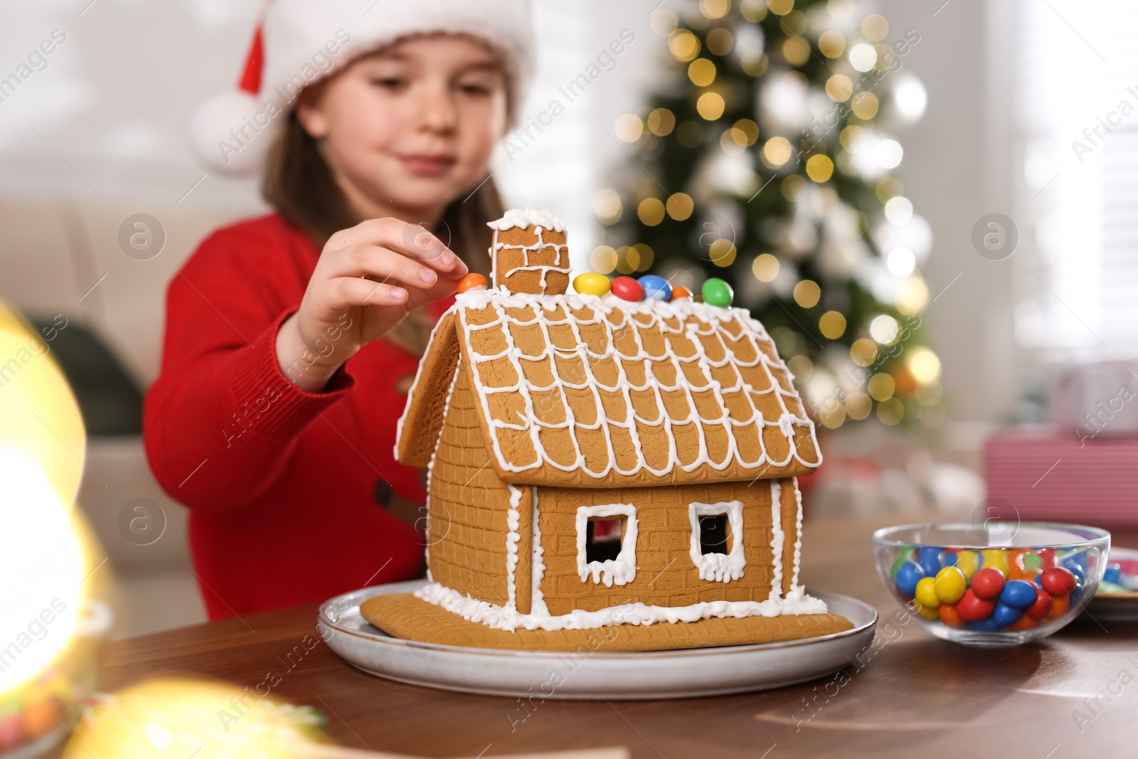 Photo of Cute little girl decorating gingerbread house at table indoors