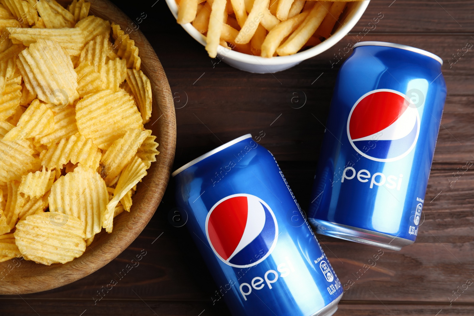 Photo of MYKOLAIV, UKRAINE - FEBRUARY 15, 2021: Cans of Pepsi and snacks on wooden table, flat lay