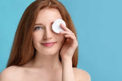 Photo of Beautiful woman with freckles covering eye with cotton pad on light blue background. Space for text