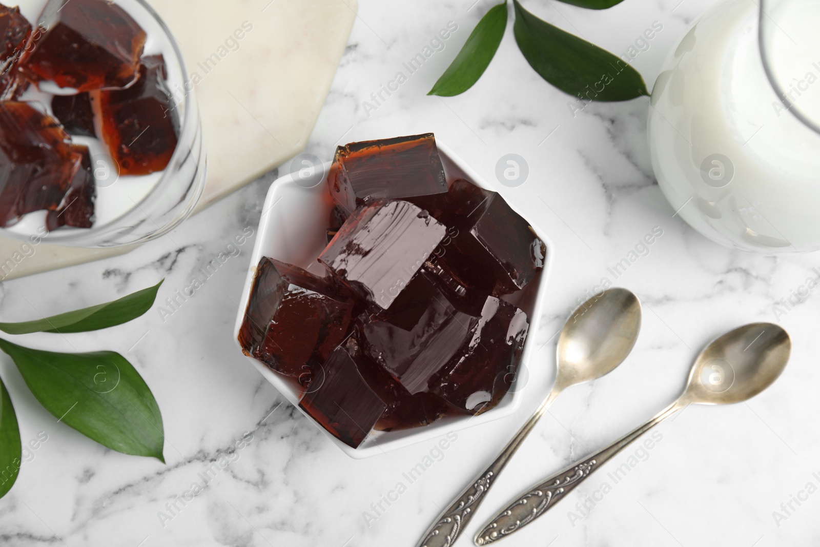 Photo of Delicious grass jelly cubes, milk and green leaves on white marble table, flat lay