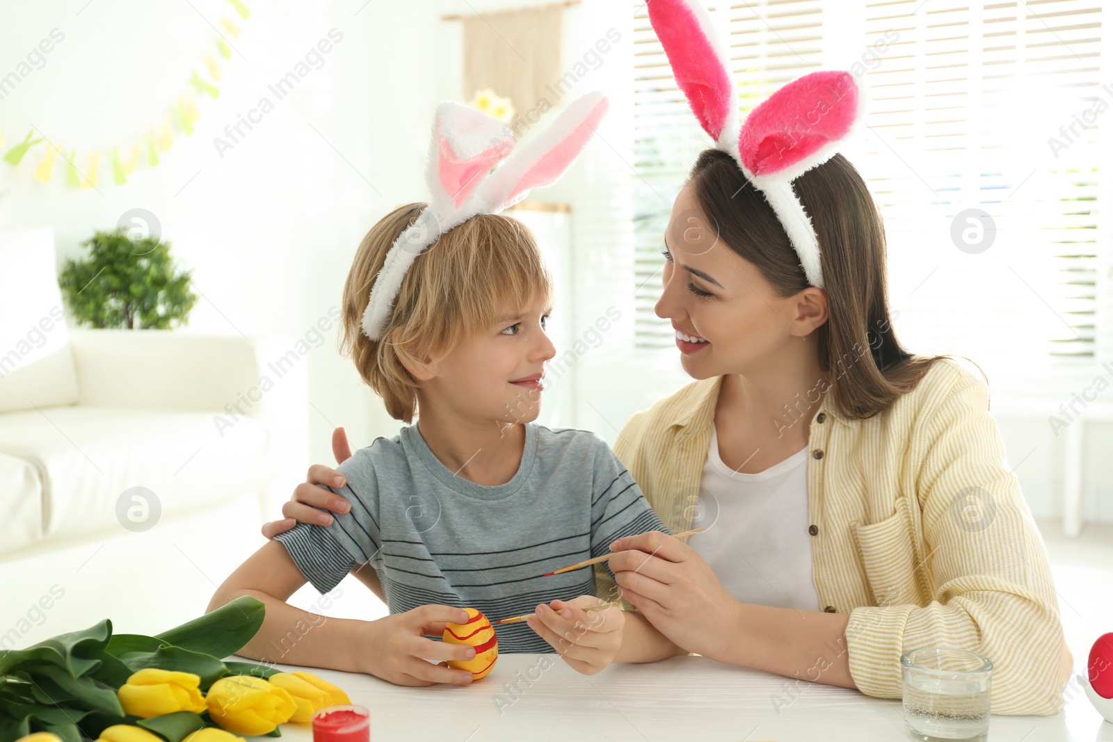 Photo of Happy mother and son with bunny ears headbands painting Easter egg at home