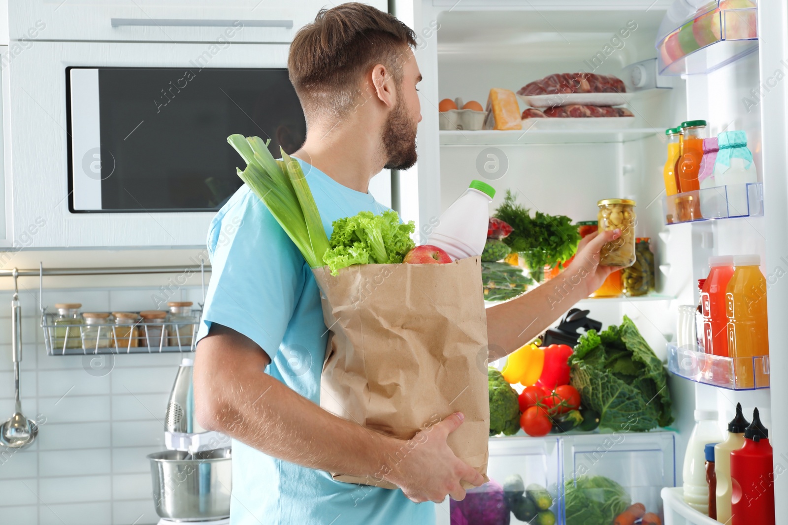 Photo of Man with paper bag putting products into refrigerator in kitchen