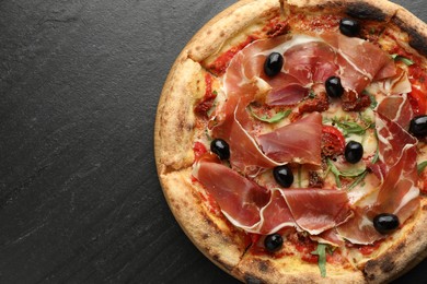 Photo of Tasty pizza with cured ham, olives and sun-dried tomato on black table, top view. Space for text