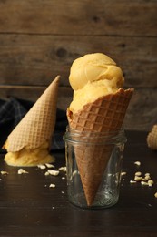 Photo of Delicious yellow ice cream in wafer cone and glass jar on black wooden table, closeup
