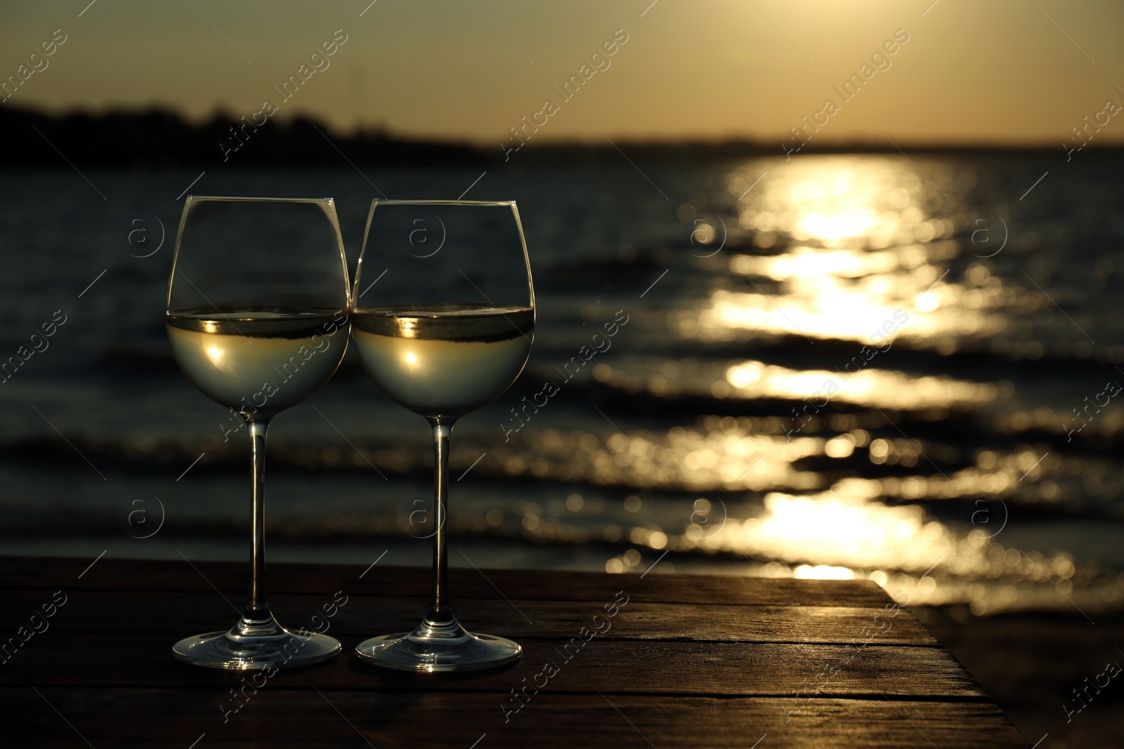 Photo of Two glasses of wine on wooden table near river at sunset. Space for text