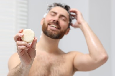 Photo of Happy man showing solid shampoo bar in bathroom, selective focus