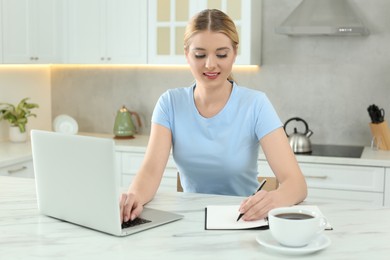 Photo of Home workplace. Woman writing in notebook near laptop at marble desk in kitchen