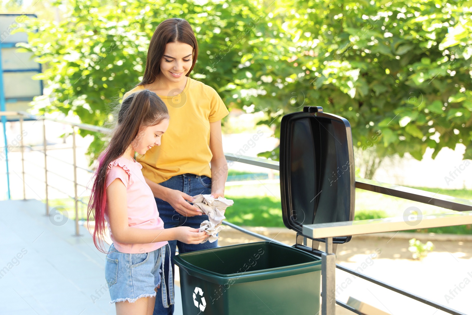 Photo of Mother and her daughter throwing paper into recycling bin outdoors