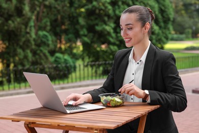 Photo of Happy businesswoman with container of salad using laptop while having lunch at wooden table outdoors