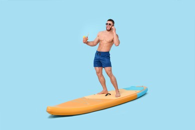 Photo of Happy man with refreshing drink talking on smartphone on SUP board against light blue background