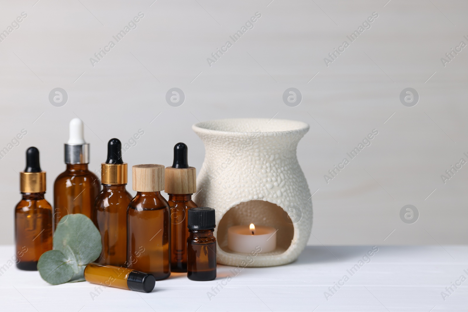 Photo of Different aromatherapy products, burning candle and eucalyptus leaves on white wooden table against light background, space for text