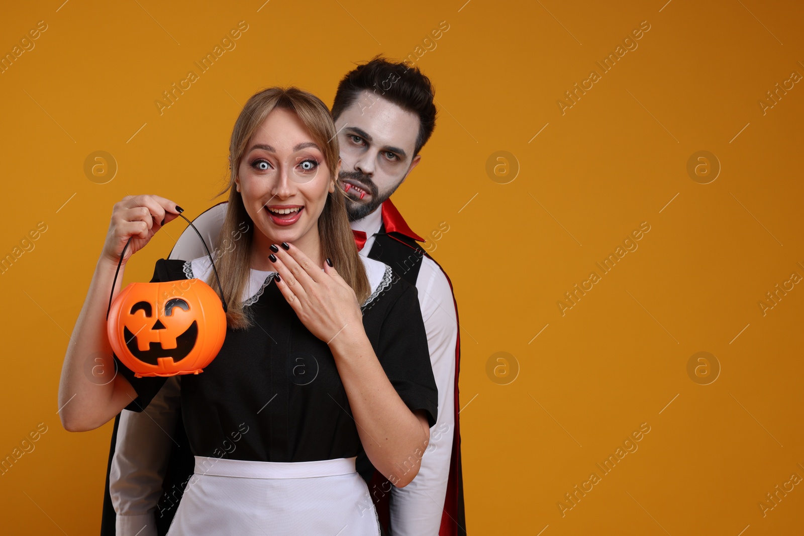 Photo of Couple in scary maid and vampire costumes with pumpkin bucket on orange background, space for text. Halloween celebration