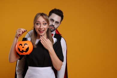 Couple in scary maid and vampire costumes with pumpkin bucket on orange background, space for text. Halloween celebration