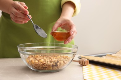 Making granola. Woman adding honey into bowl with mixture of oat flakes and other ingredients at light marble table in kitchen, closeup